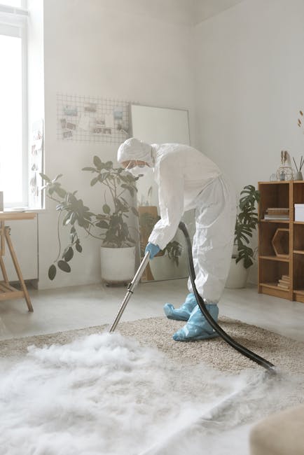 A professional cleaning technician dressed in a full white protective suit, face mask, and blue gloves is using a vacuum cleaner with a long hose to perform deep cleaning and sanitisation of a beige, 